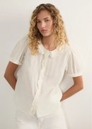 A woman with curly blonde hair wearing a white short-sleeve blouse with a ruffled Peter Pan collar and pleated front, paired with white pants, photographed against a plain light background.