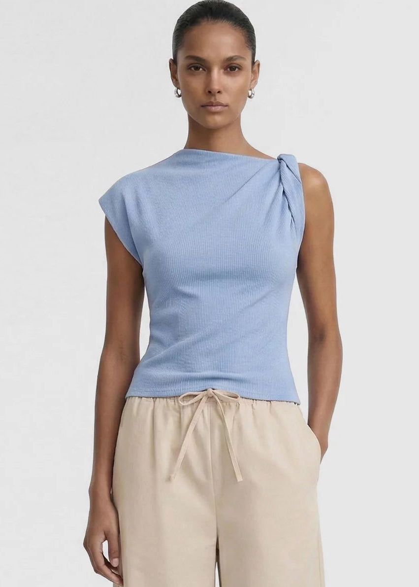 A model wearing a light blue knit top with a unique asymmetrical shoulder tie detail and beige drawstring trousers, shot against a plain light grey background.
