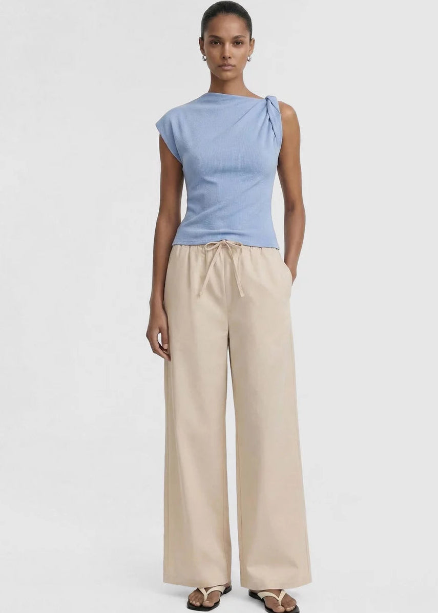 A model wearing a light blue knit top with a unique asymmetrical shoulder tie detail and beige drawstring trousers, shot against a plain light grey background.