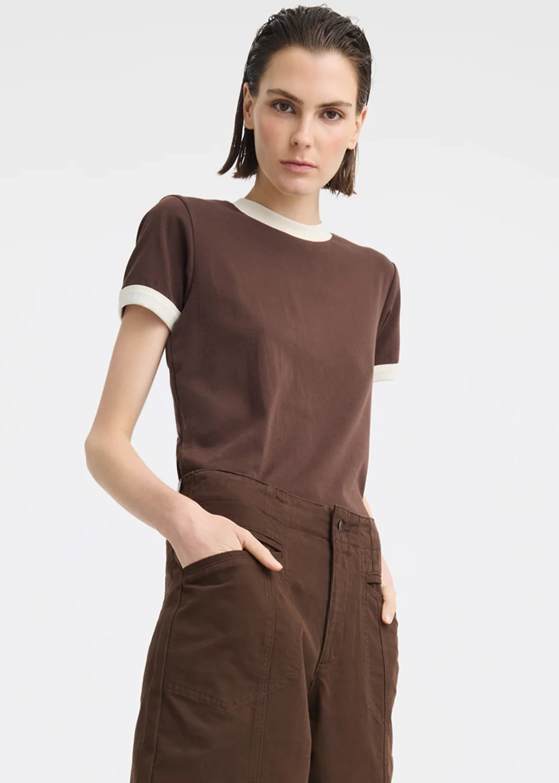 A model wearing a brown short-sleeve t-shirt with cream ribbed trim at the crew neck and sleeve cuffs, paired with brown wide-leg trousers, against a light grey studio background.