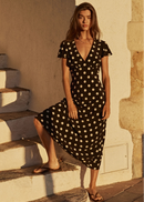 A woman wearing a black midi dress with white polka dots stands on outdoor stone steps against a sunlit beige wall.