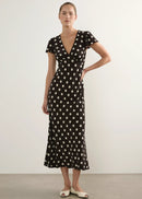 A woman modelling a black midi dress with a white polka dot pattern, short flutter sleeves, and a V-neckline, standing against a plain light grey studio background.