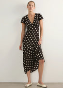 A woman modelling a black midi wrap dress with white polka dots, featuring short sleeves, a V-neckline, and an asymmetrical wrap skirt, paired with cream-colored flat shoes against a neutral studio background.