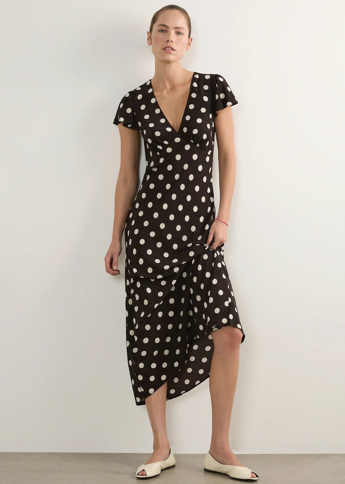 A woman modelling a black midi wrap dress with white polka dots, featuring short sleeves, a V-neckline, and an asymmetrical wrap skirt, paired with cream-colored flat shoes against a neutral studio background.