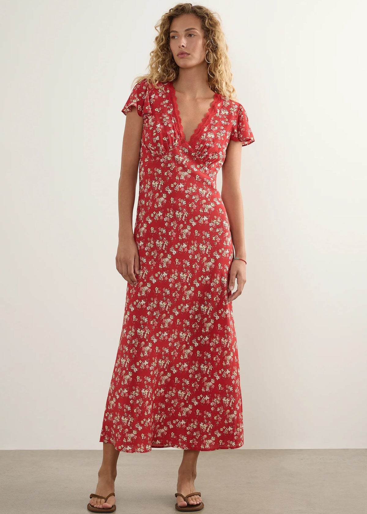 A woman modelling a red midi-length dress with a white floral pattern, short flutter sleeves, and a V-neckline, standing against a plain off-white studio wall.
