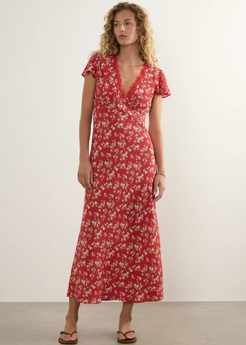 A woman modelling a red midi-length dress with a white floral pattern, short flutter sleeves, and a V-neckline, standing against a plain off-white studio wall.