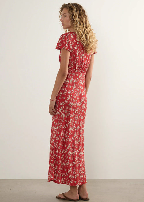 A woman with curly blonde hair is shown from the back wearing a red short-sleeve maxi dress with a white floral pattern, standing against a plain off-white wall.