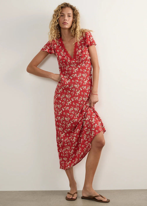A woman modelling a red midi-length dress with a white floral pattern, short flutter sleeves, and a V-neckline, standing against a plain off-white studio wall.