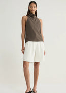 A model stands against a light grey studio background wearing a sleeveless cowl-neck top in taupe-brown and white shorts, paired with black strappy sandals.