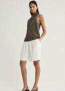 A model stands against a light grey studio background wearing a sleeveless, mock-neck, dark brown knit top and white shorts, paired with black wedge sandals.