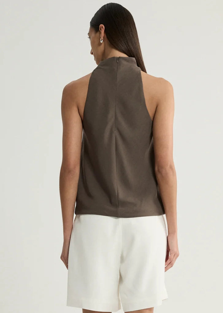 A woman shown from the back wearing a sleeveless taupe-brown top and white shorts against a plain light grey studio background.