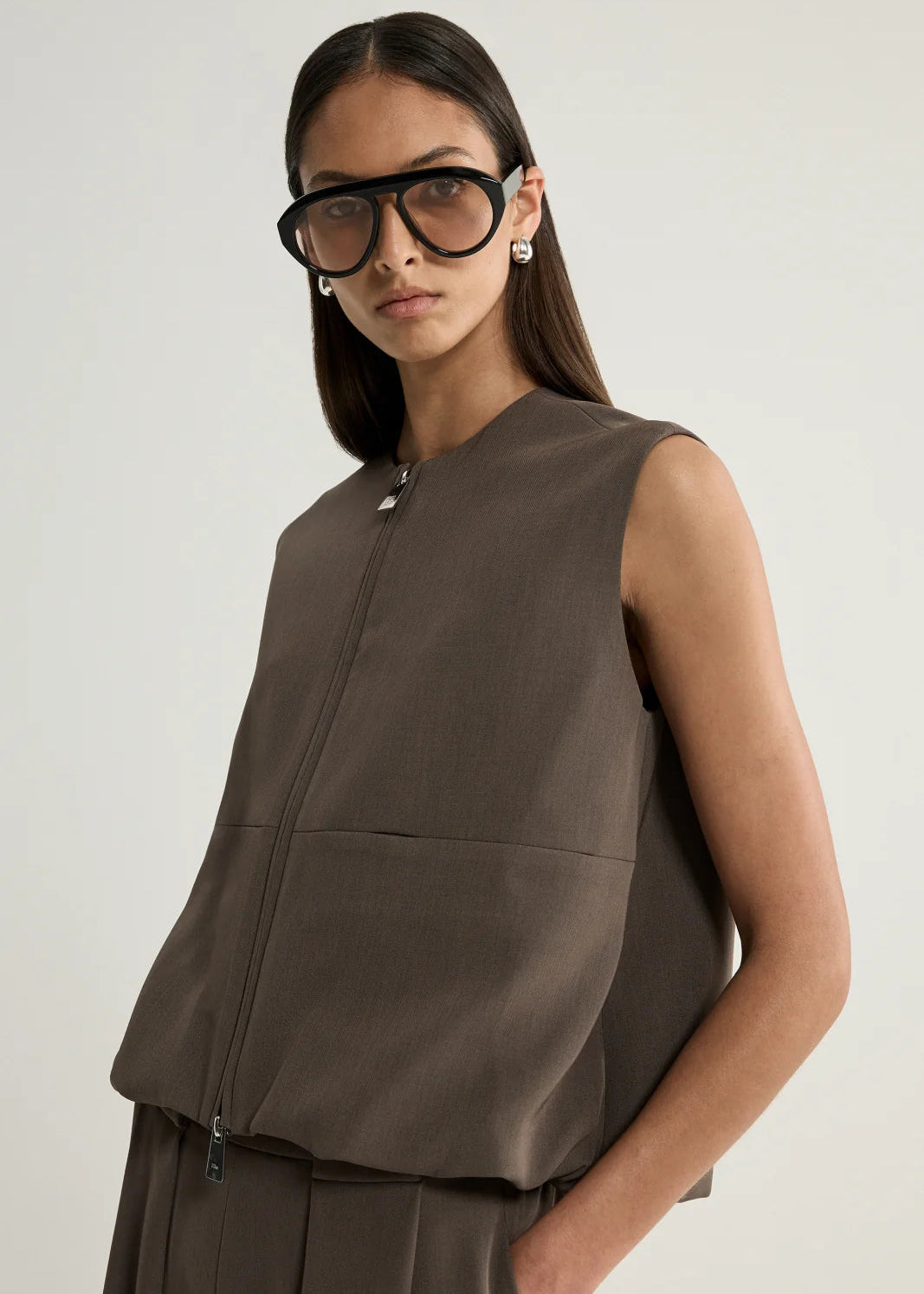 A model wearing a sleeveless taupe-brown zip-front vest and matching trousers, styled with oversized black sunglasses, against a light grey studio background.