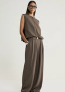 A woman modelling a sleeveless taupe-brown jumpsuit with a front zipper and wide-leg pants, styled with black sandals and glasses against a light grey studio background.