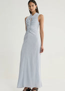 A woman modelling a sleeveless light blue-grey maxi dress with a gathered ruched bodice and keyhole neckline, paired with black wedge sandals, against a plain light grey studio background.