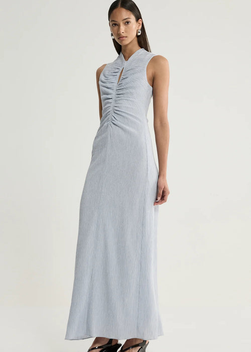 A woman modelling a sleeveless light blue-grey maxi dress with a gathered ruched bodice and keyhole neckline, paired with black wedge sandals, against a plain light grey studio background.