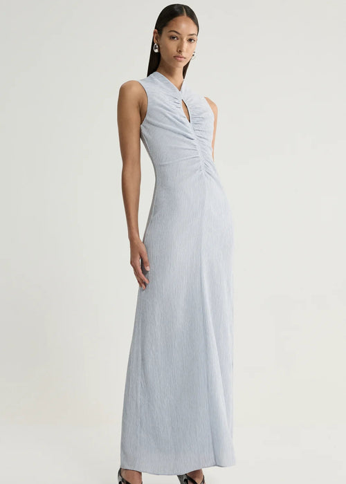 A woman modelling a sleeveless light blue-grey maxi dress with a gathered ruched bodice and keyhole neckline, paired with black wedge sandals, against a plain light grey studio background.