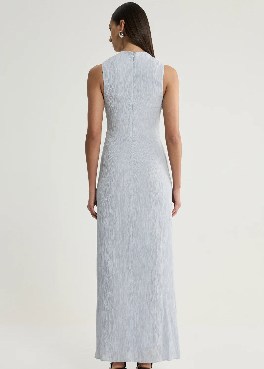 A woman viewed from the back wearing a sleeveless, floor-length light blue dress against a plain light grey studio background.