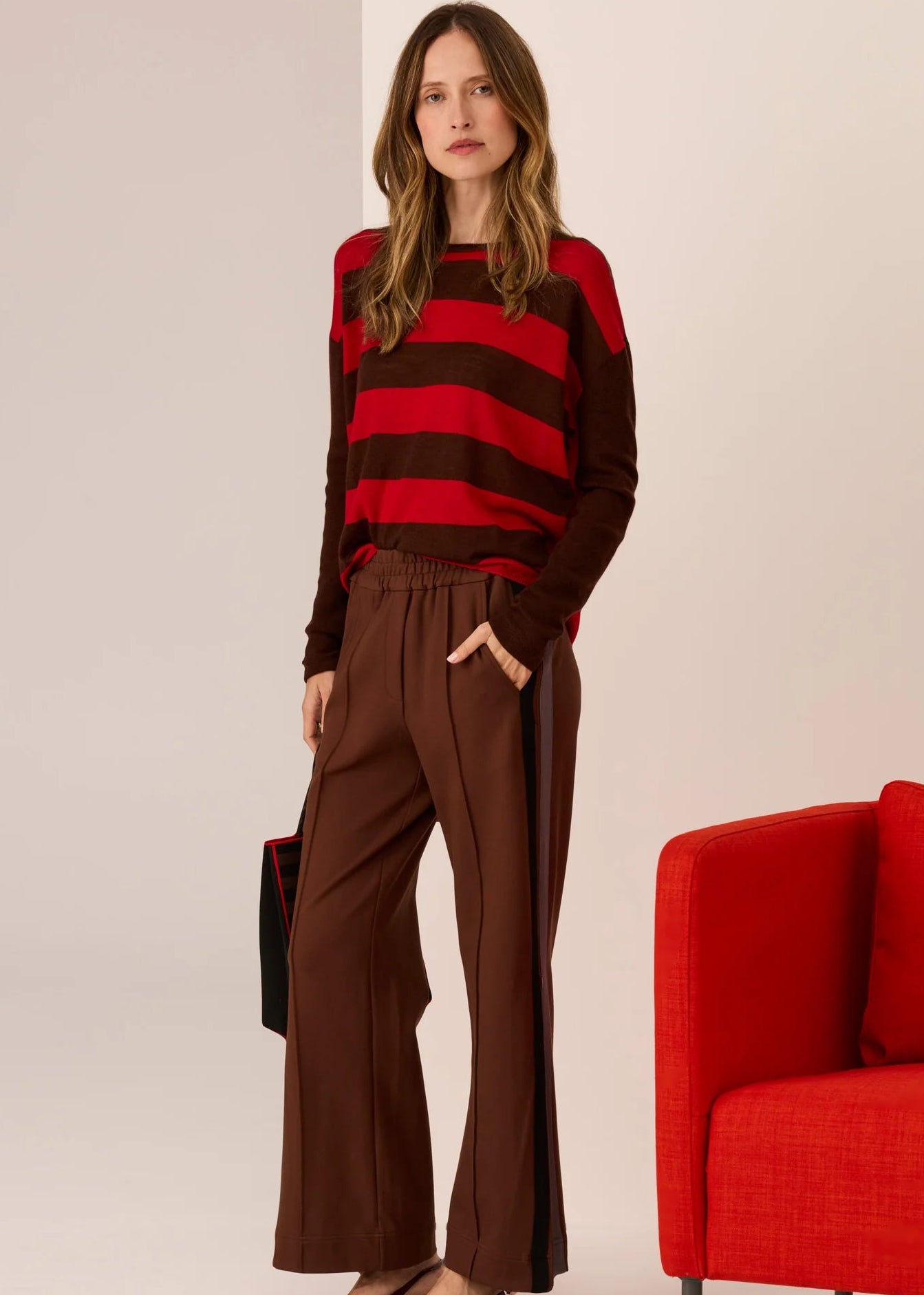 Woman wearing a red and brown striped sweater with brown pants, standing next to a red chair on a beige background.