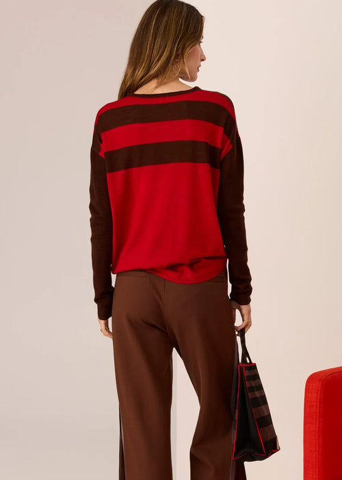 Woman wearing a red and brown striped sweater with brown pants, standing next to a red chair on a beige background.
