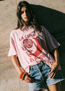 A woman wearing a light pink oversized graphic T-shirt with a red rose and crescent moon design and the text 'LA LUNA ELANOR', paired with denim shorts, leaning against a textured concrete wall.