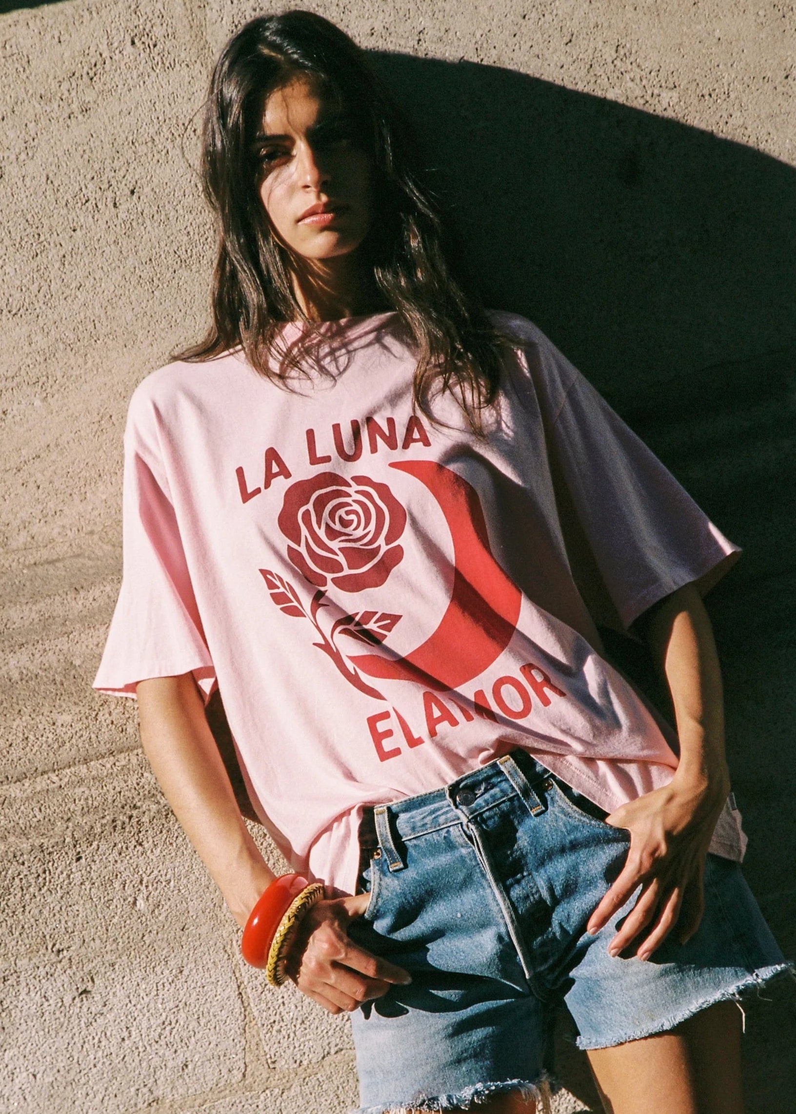 A woman wearing a light pink oversized graphic T-shirt with a red rose and crescent moon design and the text 'LA LUNA ELANOR', paired with denim shorts, leaning against a textured concrete wall.