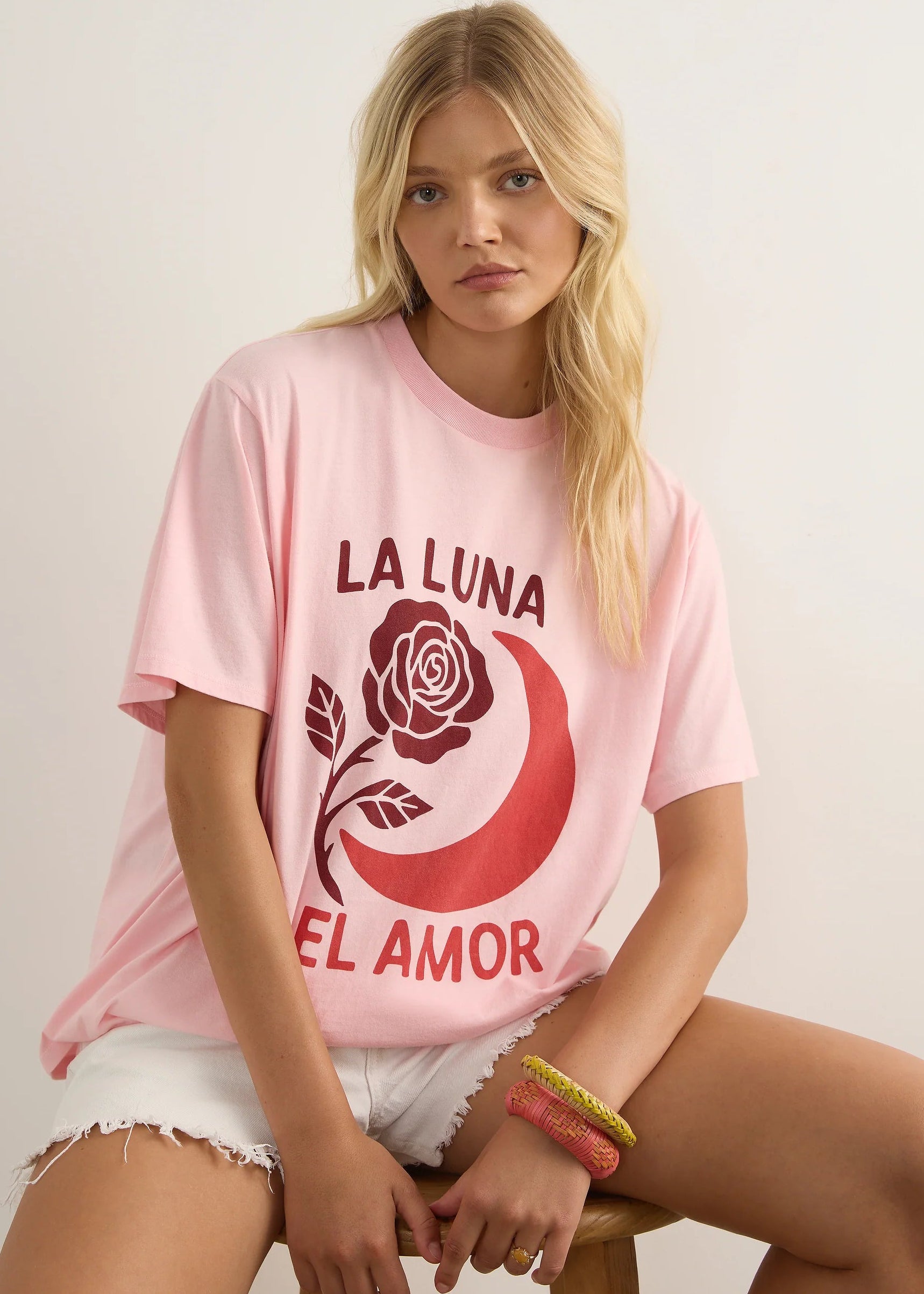 A blonde woman sits on a wooden stool wearing an oversized light pink graphic T-shirt with a red crescent moon, rose, and the text 'LA LUNA EL AMOR'.