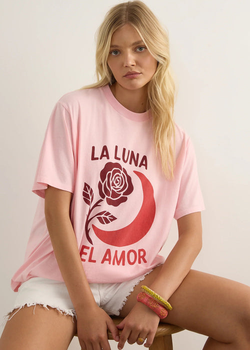 A blonde woman sits on a wooden stool wearing an oversized light pink graphic T-shirt with a red crescent moon, rose, and the text 'LA LUNA EL AMOR'.