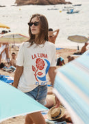 A woman wearing a white graphic t-shirt with a red rose and blue crescent design, standing on a sandy beach with the ocean and umbrellas in the background.
