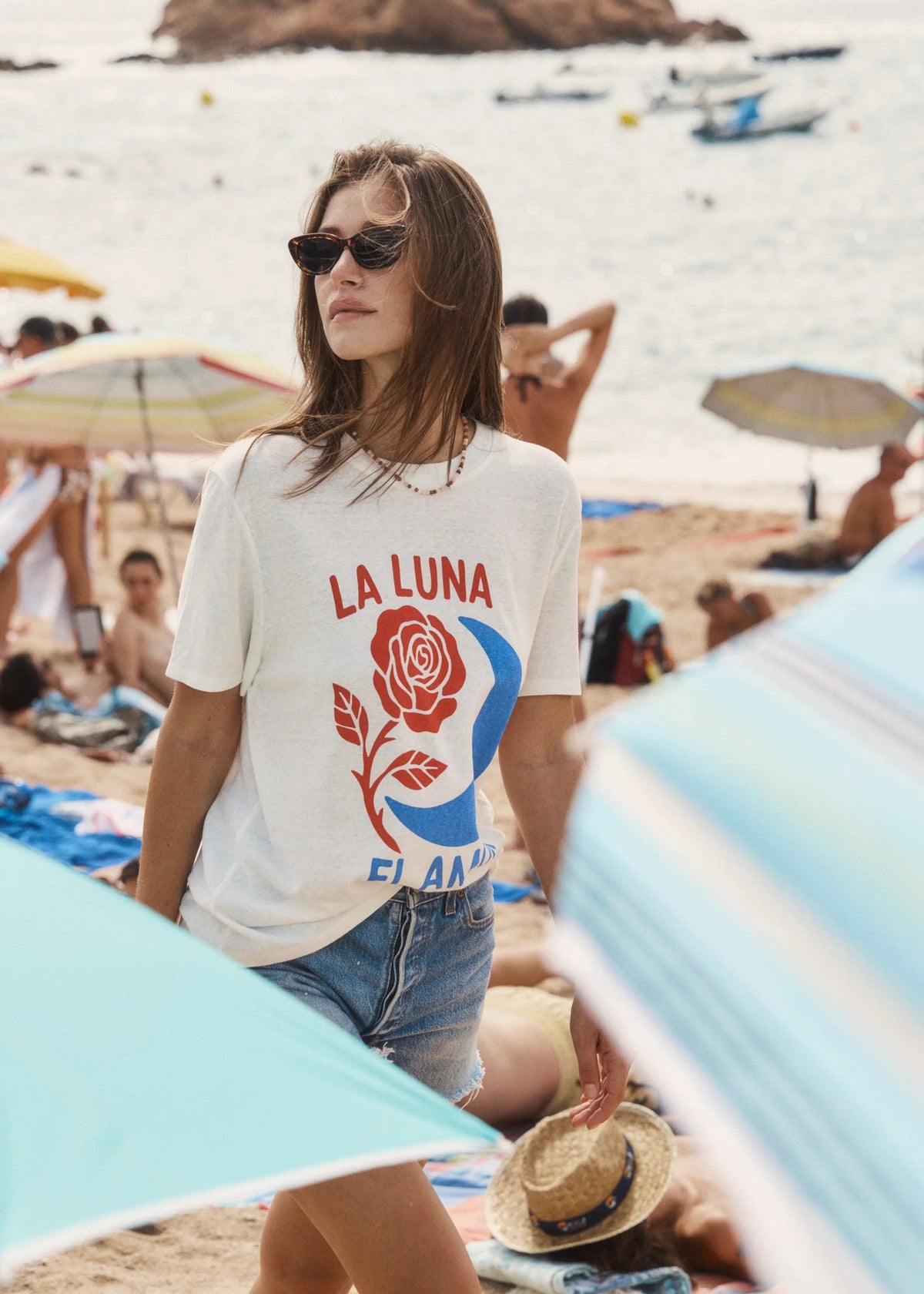 A woman wearing a white graphic t-shirt with a red rose and blue crescent design, standing on a sandy beach with the ocean and umbrellas in the background.