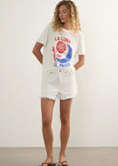 A woman with curly blonde hair wearing a white graphic t-shirt featuring a red rose, blue crescent moon, and the Spanish text 'LA LUNA EL AMOR' in red and blue, paired with white denim shorts against a plain light background.