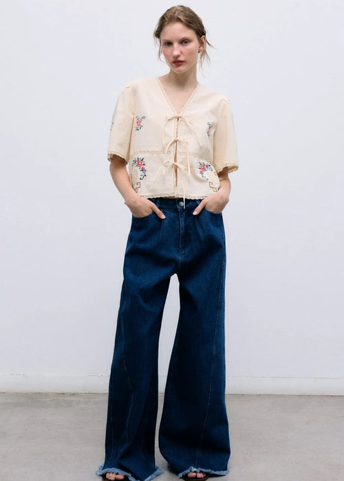 Woman wearing a beige embroidered top and blue wide-leg pants against a white background