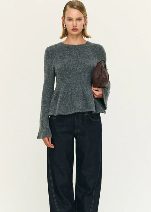 Woman wearing a grey top and dark pants holding a brown clutch against a white background