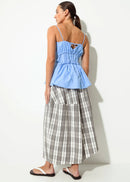 A woman stands in profile wearing a light blue sleeveless peplum-style top with thin straps and a grey-and-white plaid midi skirt, paired with black-and-white sandals, against a plain off-white wall.