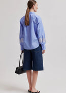 Woman wearing a blue striped shirt and dark shorts, holding a black handbag on a light grey background