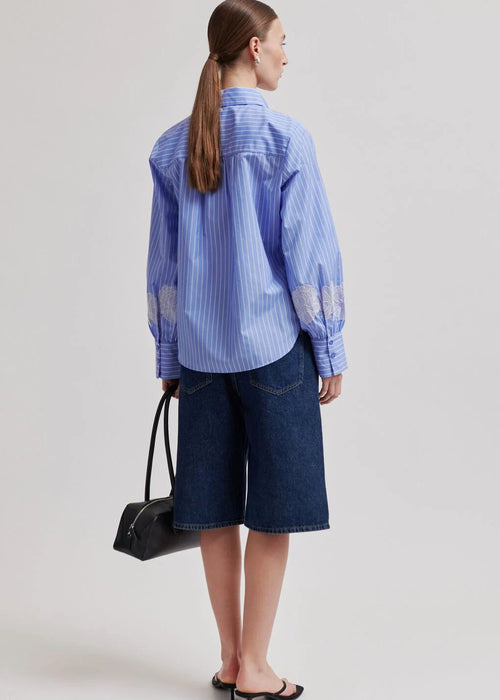 Woman wearing a blue striped shirt and dark shorts, holding a black handbag on a light grey background