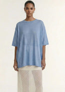 A model stands against a plain light beige studio background wearing a loose light blue short-sleeve top and a sheer cream maxi skirt with small white polka dots.