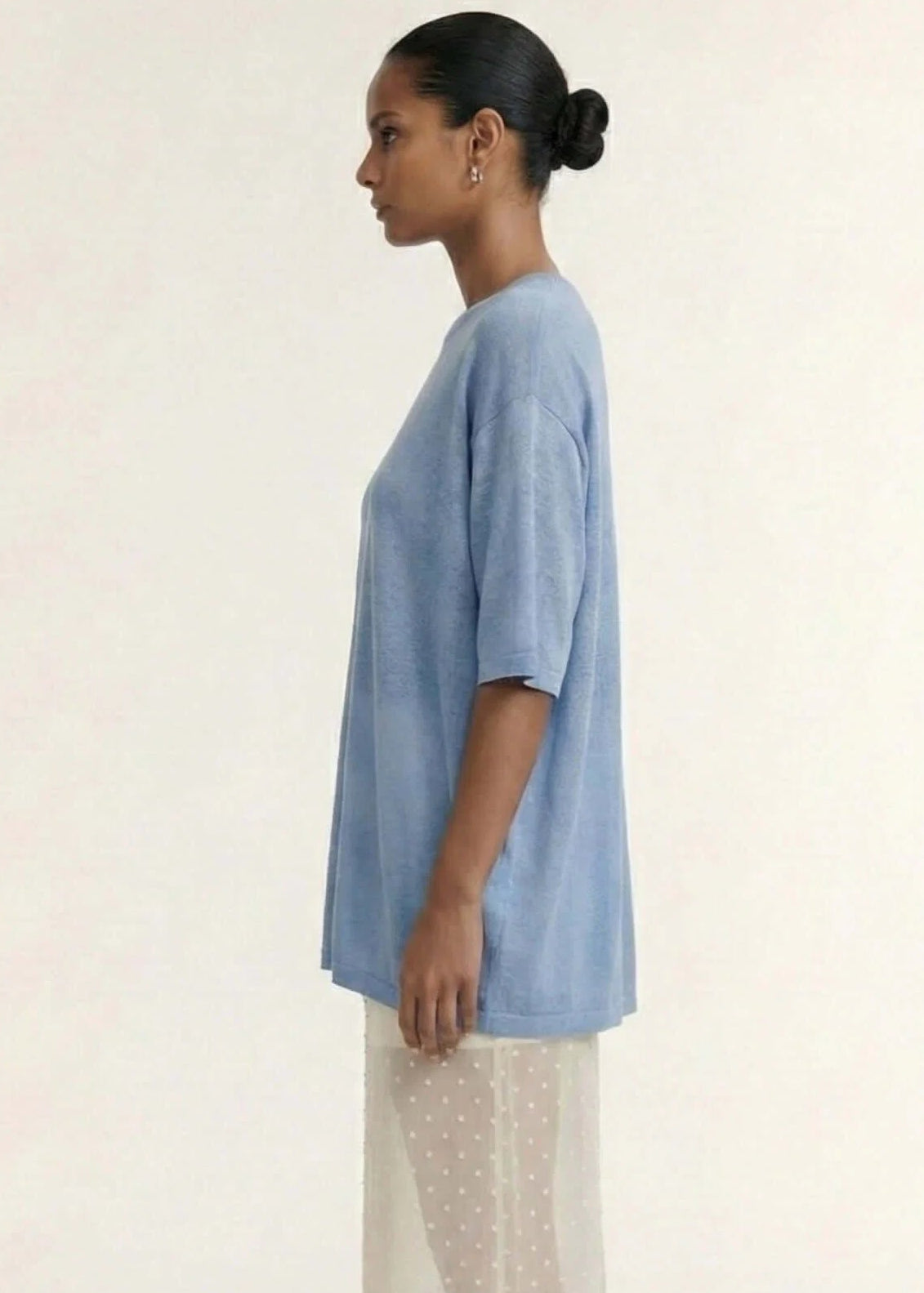 A model stands against a plain light beige studio background wearing a loose light blue short-sleeve top and a sheer cream maxi skirt with small white polka dots.