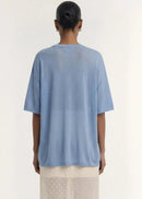 A model stands against a plain light beige studio background wearing a loose light blue short-sleeve top and a sheer cream maxi skirt with small white polka dots.