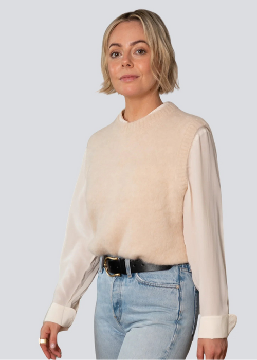 Woman wearing a beige sweater over a white shirt with light blue jeans on a plain background