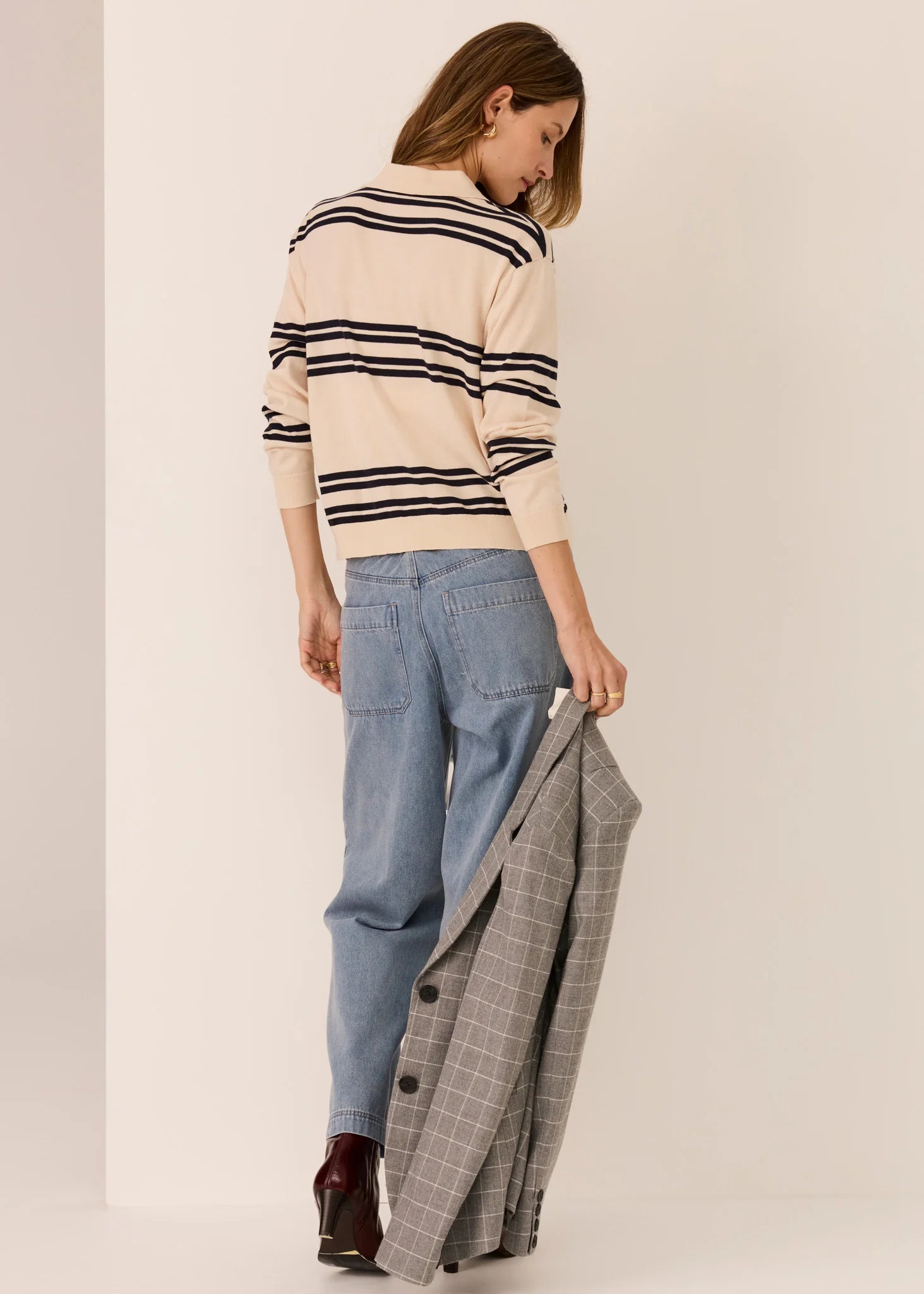 Woman wearing a striped sweater and jeans, holding a plaid jacket, on a plain background