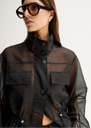 A model wearing a black sheer long-sleeve button-up shirt with a high collar and a matching black mini skirt, styled with black sunglasses and flat sandals, against a plain white studio background.