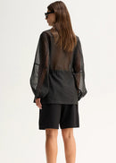 A woman shown from the back wearing a sheer black long-sleeve top with a gathered waist and black shorts, standing against a plain light background.