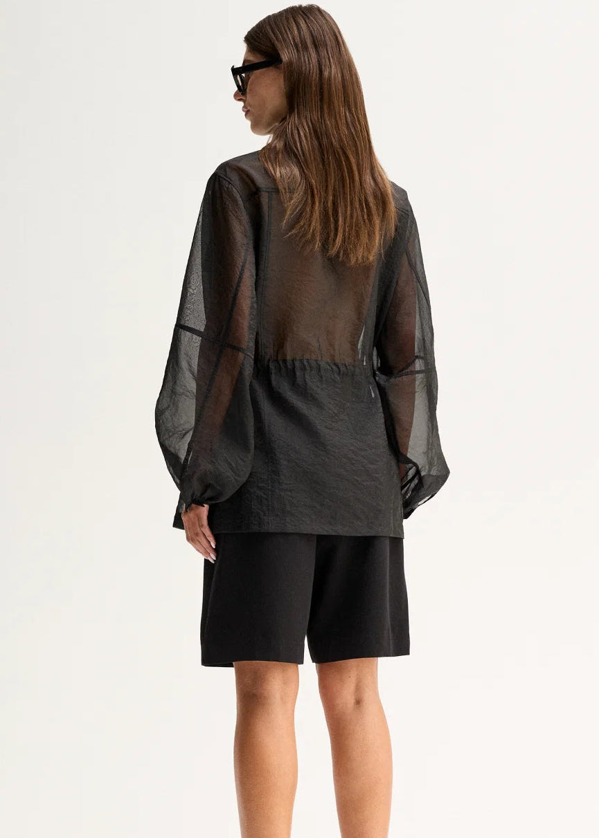 A woman shown from the back wearing a sheer black long-sleeve top with a gathered waist and black shorts, standing against a plain light background.
