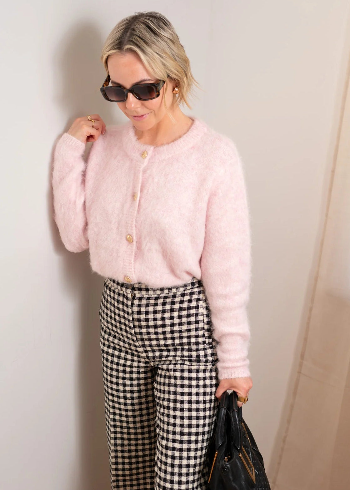Woman wearing a pink cardigan and checkered pants with sunglasses on a light grey background