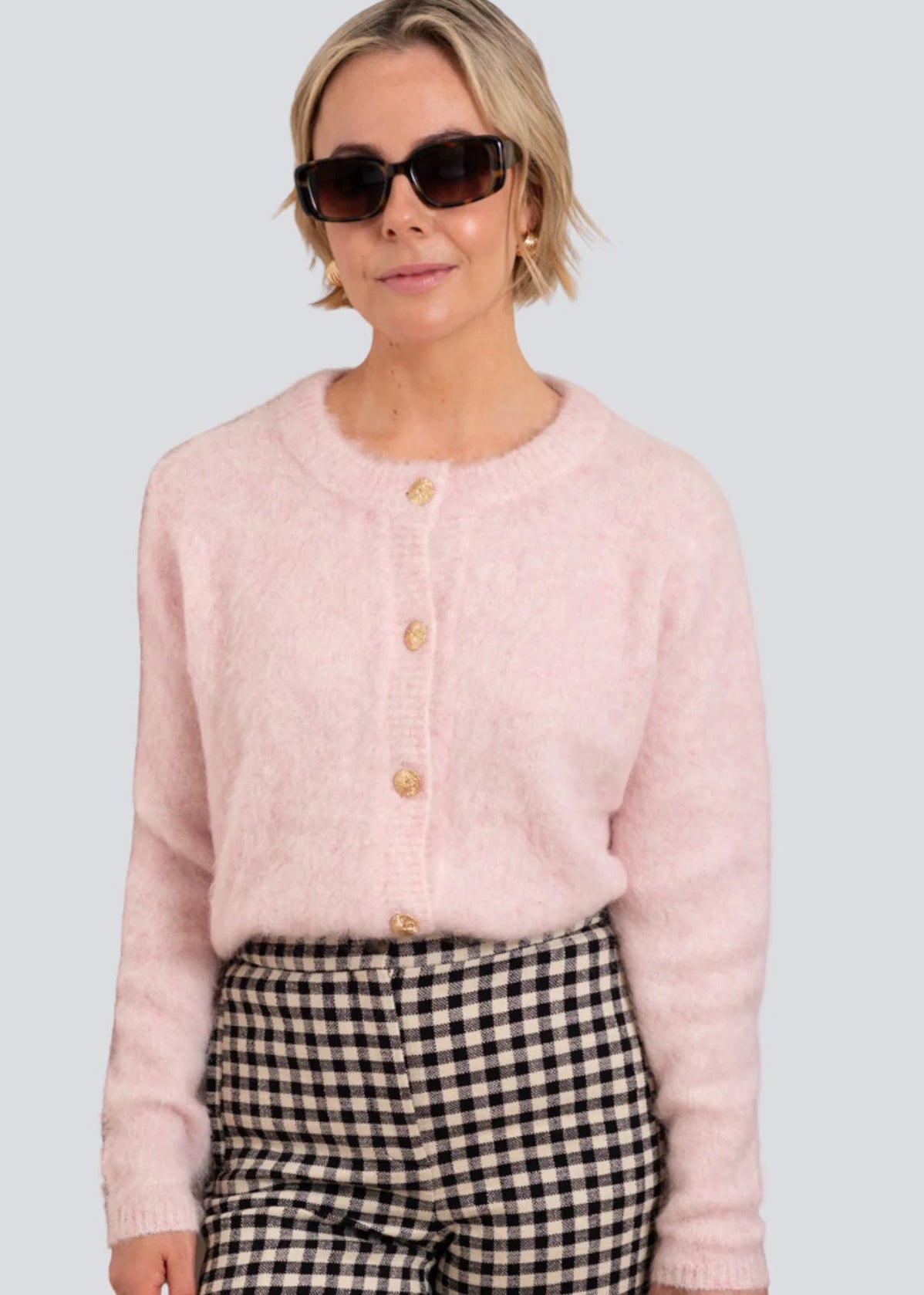 Woman wearing a pink cardigan and checkered pants with sunglasses on a light grey background