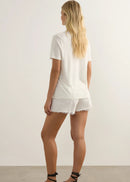 A woman modelling a white short-sleeve graphic T-shirt with 'RADICAL LOVE' printed in black on the chest, paired with white frayed denim shorts and black strappy sandals, standing against a plain light grey studio background.
