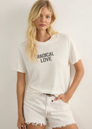 A woman modelling a white short-sleeve graphic T-shirt with 'RADICAL LOVE' printed in black on the chest, paired with white frayed denim shorts and black strappy sandals, standing against a plain light grey studio background.