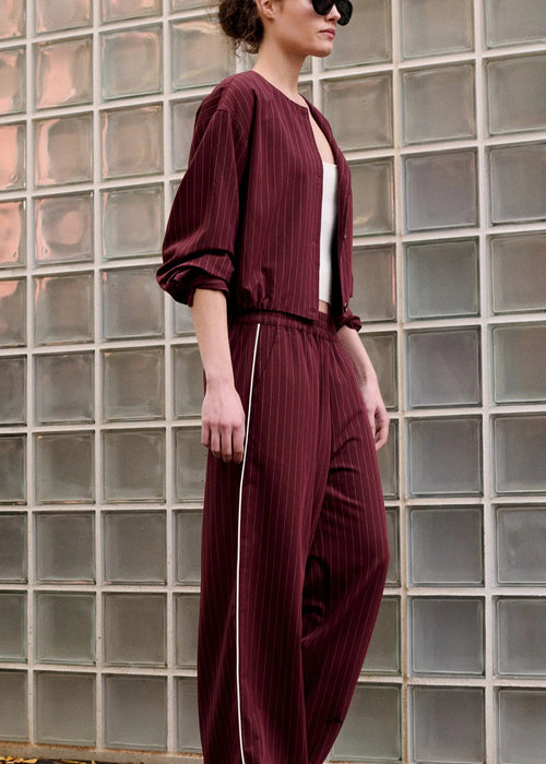 A woman wearing a pleated burgundy jumpsuit with a white inner top and white side piping, standing in front of a glass block wall.