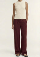 A model stands against a plain light background wearing a sleeveless cream ribbed knit top and dark burgundy pleated wide-leg trousers with gold-tone sandals.