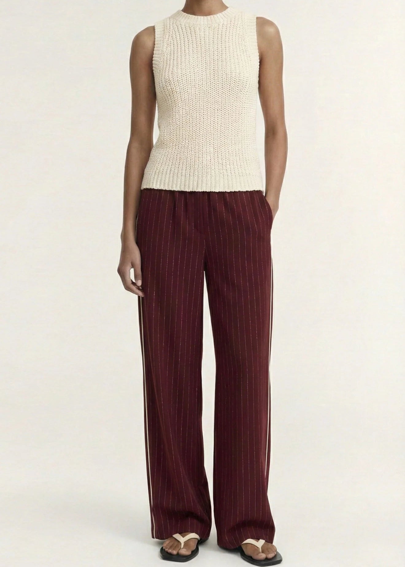 A model stands against a plain light background wearing a sleeveless cream ribbed knit top and dark burgundy pleated wide-leg trousers with gold-tone sandals.
